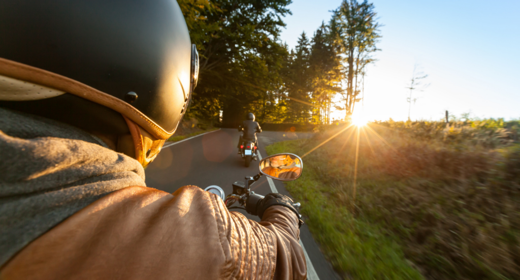 Primer plano en primera persona de un motorista rodando por una carretera virada entre pinos al atardecer, ideal para ilustrar la ruta de montaña de Valsaín.